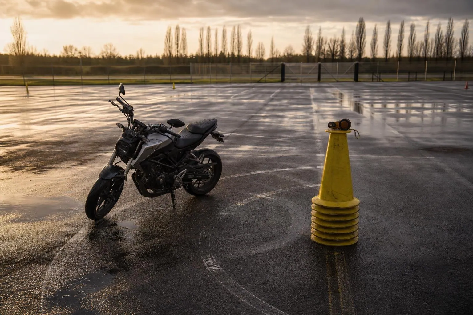 Une moto école seule sur une piste plateau mouillée en fin de journée, à côté d’une pile de plots jaunes rangés