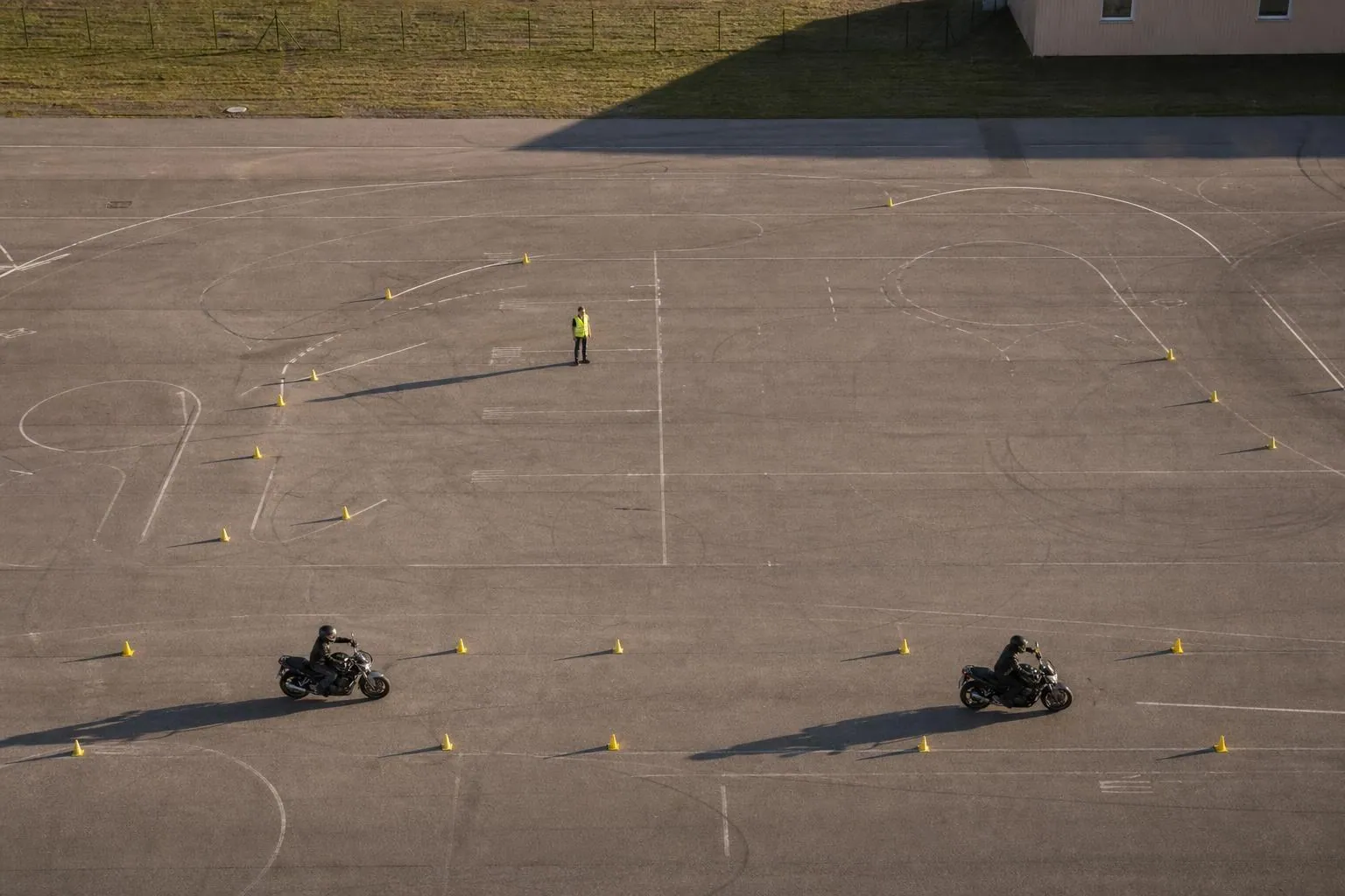 Vue en plongée d’une piste plateau où deux motos école tracent des trajectoires entre des paires de plots jaunes