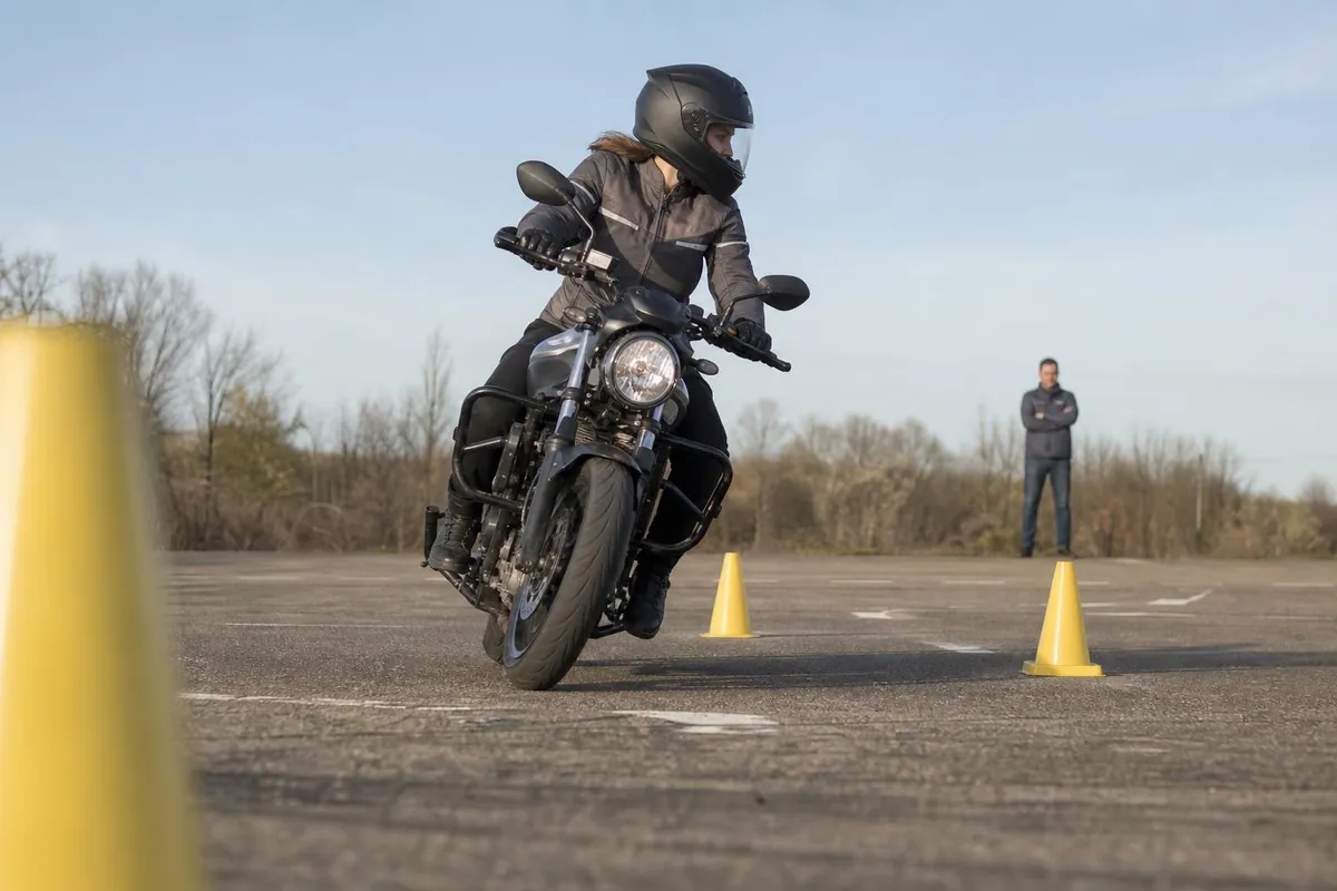 Élève casquée penchée sur une moto à faible allure entre deux plots jaunes d’une piste plateau, les deux pieds sur les repose pieds, l’enseignant en observation à l’arrière plan